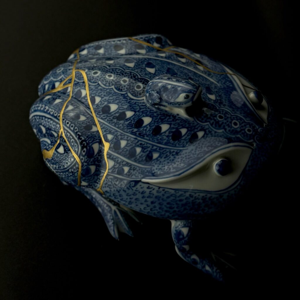 Blau-weiße Porzellandose in Froschform mit goldenen Kintsugi-Linien auf dem Rücken, vor schwarzem Hintergrund