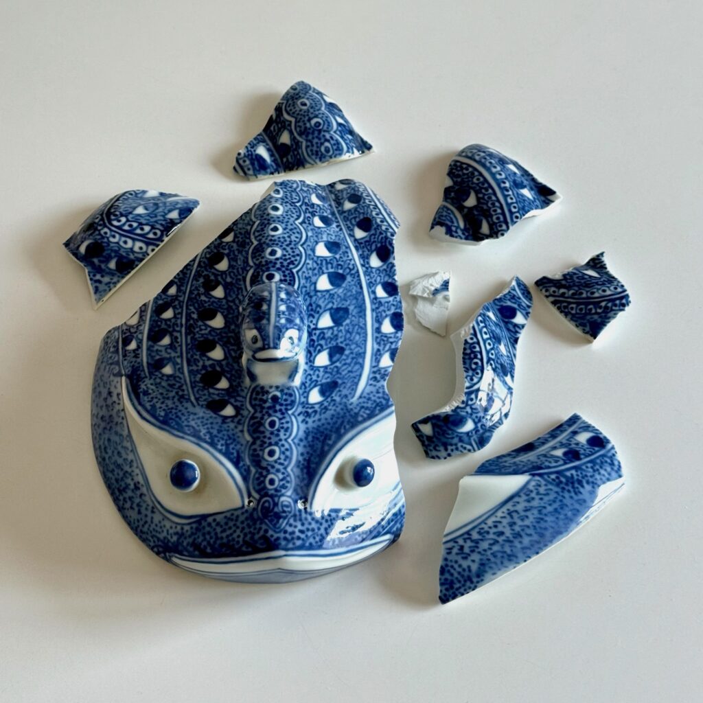 Alt: Zerbrochener Froschdeckel, ein größeres Stück mit Knauffrosch und sieben einzelne Scherben auf weißem Grund